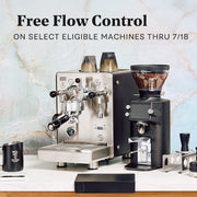 Promotional product photo showing a stainless steel espresso machine and a black coffee grinder on a countertop, with text reading 'Free Flow Control ON SELECT ELIGIBLE MACHINES THRU 7/15' above them. A marble-pattern backdrop and a few small accessories are visible around the machines.