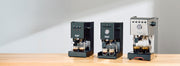 Gaggia Classic Pro E24, Gaggia Classic UP and Gaggia Classic GT next to each other on a wooden counter top in front of a gray wall.