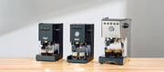 Close-up of the Gaggia Classic Pro E24, Gaggia Classic UP and Gaggia Classic GT next to each other on a wooden counter top in front of a gray wall.
