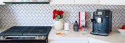 Gaggia Cadorna Milk in Black on a white kitchen counter next to a bag of coffee, chocolate sauce, flowers and a stove.