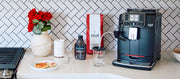 Gaggia Cadorna Milk in Black on a white kitchen counter next to a bag of coffee, chocolate sauce, flowers and a stove.