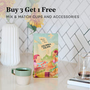 A bag of Whole Latte Love Colombia Tolima coffee stands on a countertop in a styled kitchen scene, with a mug, glass carafe, and floral decor in the background. Large text at the top reads 'Buy 3 Get 1 Free' and 'MIX & MATCH CUPS AND ACCESSORIES'.