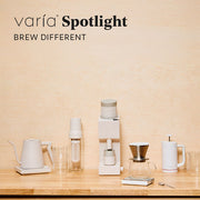A collection of Varia products in white and clear arranged on a light wood surface against a beige wall, with the text 'Varía Spotlight BREW DIFFERENT'.