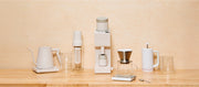 Close-up of a collection white and clear Varia products on a wooden counter top in front of a beige background.