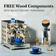 Close-up of a Rocket Appartamento TCA Espresso Machine with Copper Panels and Olive Wood Accents next to a Eureka Oro Mignon Grinder in White and a Blue notNeutral  latte cup on a gray stone-textured surface against a light gray wall. Promotional text overlayed that reads "FREE Wood Components with select rocket purchases".