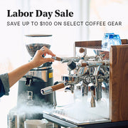 A person's hand is shown adjusting the steam wand on a ECM Synchronika with wooden accents and a Ceado coffee grinder in the background with steam rising. Large text at the top reads 'Labor Day Sale SAVE UP TO $100 ON SELECT COFFEE GEAR'.