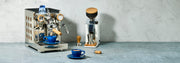 Rocket Appartamento TCA Espresso Machine with Copper Panels and Olive Wood Accents next to a Eureka Oro Mignon Grinder in White and a Blue notNeutral  latte cup on a gray stone-textured surface against a light gray wall.