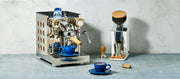 Close-up of a Rocket Appartamento TCA Espresso Machine with Copper Panels and Olive Wood Accents next to a Eureka Oro Mignon Grinder in White and a Blue notNeutral  latte cup on a gray stone-textured surface against a light gray wall.