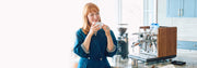 Woman holding a coffee cup in a kitchen with an ECM Synchronika Espresso Machine with wood accents next to a Ceado Grinder on a marble counter. 
