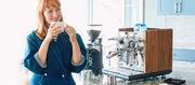 Close-up of a woman holding a coffee cup in a kitchen with an ECM Synchronika Espresso Machine with wood accents next to a Ceado Grinder on a marble counter. 