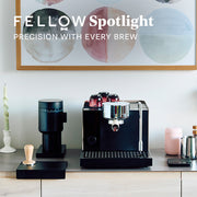 A Fellow Espresso Series 1 next to a Fellow Opus Grinder on a counter top next to coffee accessories. Promotional text overlayed that reads "Fellow Spotlight - Precision with every brew".