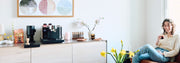 A woman sitting in a chair besides a Fellow Espresso Series 1 next to a Fellow Opus Grinder on a counter top next to coffee accessories, Whole latte Love Hidden Malabar coffee and a flower decorations.
