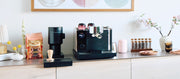 Close-up of a Fellow Espresso Series 1 next to a Fellow Opus Grinder on a counter top next to coffee accessories, Whole latte Love Hidden Malabar coffee and a flower decorations.