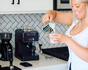 A woman in a white tank top pours milk from a stainless steel pitcher into a cup on a kitchen counter, with a black espresso machine and coffee grinder behind her.