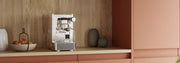 Stone Lite Espresso Machine in stainless steel on a wooden counter top with salmon colored cupboards next to a bowl of apples in front of a wooden wall.