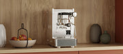 Close-up of a Stone Lite Espresso Machine in stainless steel on a wooden counter top with salmon colored cupboards next to a bowl of apples in front of a wooden wall.