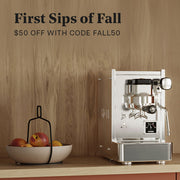 Stone Lite Espresso Machine in stainless steel on a wooden counter top next to a bowl of apples in front of a wooden wall. Promotional text overlayed that reads "First Sips of Fall - $50 OFF with code FALL50".