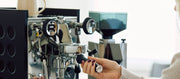 Close-up of a person holding the portafilter on a Rocket Espresso Appartamento in Anthracite with black panels and a grinder in the background.