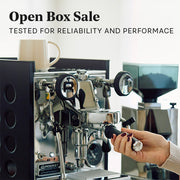 Close-up of a person holding the portafilter on a Rocket Espresso Appartamento in Anthracite with black panels and a grinder in the background. Promotional text overlayed that reads "Open Box Sale - Tested for reliability and performance".