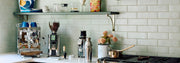 A styled kitchen countertop featuring an ECM Mechanika Max II in stainless steel, an ECM Exacto Grinder, an ECM C-Automatik 54 On-Demand Espresso Grinder a metal shaker, and a small copper pot, set against a sage subway tile backsplash with a floating shelf above.