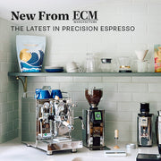 Close-up of a styled kitchen countertop featuring an ECM Mechanika Max II in stainless steel, an ECM Exacto Grinder, and a ECM C-Automatik 54 On-Demand Espresso Grinder set against a sage subway tile backsplash with a floating shelf above with Whole Latte Love Crema Wave and other coffee accessories on it. Promotional text overlayed that reads "New From ECM - The latest in precision espresso".