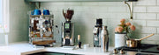 Close-up of a styled kitchen countertop featuring an ECM Mechanika Max II in stainless steel, an ECM Exacto Grinder, an ECM C-Automatik 54 On-Demand Espresso Grinder a metal shaker, and a small copper pot, set against a sage subway tile backsplash with a floating shelf above.
