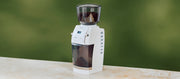 Close-up of a Baratza Vario+ in white on a beige counter top in front of a green wall.