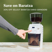 Person using a Baratza Vario+ in white on a beige counter top in front of a green wall. Promotional text overlayed that reads "Save on Baratza - 20% OFF Select Baratza Vario Grinders".