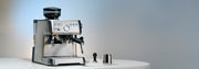Solis Grind and Infuse Perfetta in Stainless Steel on a white counter top next to a tamper and milk frothing pitcher in front of a blue and gray gradient background.
