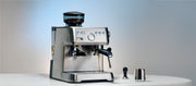 Close-up of a Solis Grind and Infuse Perfetta in Stainless Steel on a white counter top next to a tamper and milk frothing pitcher in front of a blue and gray gradient background.