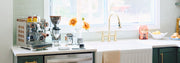 A coffee station setup on a countertop with a stainless steel Rocket Espresso Appartamento, a Rocket Espresso coffee grinder, milk pitcher and scale photographed in a bright kitchen next to a sink faucet in front of a window.