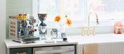Close-up of a coffee station setup on a countertop with a stainless steel Rocket Espresso Appartamento, a Rocket Espresso coffee grinder, milk pitcher and scale photographed in a bright kitchen next to a sink faucet in front of a window.