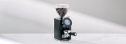 Close-up of the LX Italia Grinder in black with a portafilter on a white counter in front of a gray wall. 