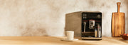 Gaggia Accademia in Black Glass next to a white mug on a brown counter top in front of a gray and black wall.