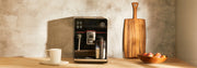 Close-up of a Gaggia Accademia in Black Glass next to a white mug on a brown counter top in front of a gray and black wall.