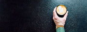 A hand holding a black cup of latte art coffee on a dark textured surface.