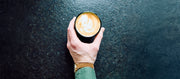Close-up of a hand holding a black cup of latte art coffee on a dark textured surface.