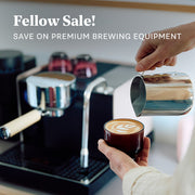 A person pouring milk from a stainless steel milk frothing pitcher into a small cup of espresso with latte art, with a Fellow Espresso Series 1 coffee machine in the background and the text 'Fellow Sale! SAVE ON PREMIUM BREWING EQUIPMENT' at the top.