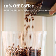 Coffee beans pouring from a dark metal canister into a clear grinder hopper, with promotional text reading '10% Off Coffee' and 'SAVE ON SELECT COFFEE WITH CODE BEANS10' overlaid at the top.