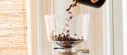 Close-up of coffee beans being poured from a black container into a clear grinder hopper, with a bamboo slat window shade in the background.