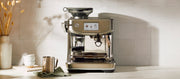 Close-up of the Breville Barista Touch Impress in Stainless Steel next to a milk frothing pitcher, cups and vase on a white counter top in front of a white tiled wall.