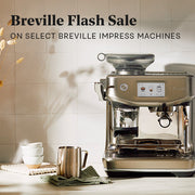 Breville Barista Touch Impress in Stainless Steel next to a milk frothing pitcher and cups on a white counter top in front of a white tiled wall. Promotional text overlayed that reads Breville Flash Sale - On Select Breville Impress Machines.
