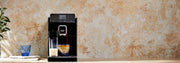 A black Gaggia Magenta Prestige and cup of coffee, shown on a countertop against a textured beige wall, with a blue glass next to it.