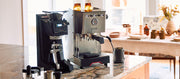 Close-up of a Etzinger etzMAX LT in Black next to a Gaggia Classic GT and other coffee accessories on a granite counter top with a fall table setting in the background.