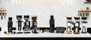 A close-up of professional coffee grinders  arranged on a white countertop against a light marble backsplash. The machines feature black, silver, and stainless steel finishes.