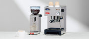 Close-up of the Lelit Anna Espresso Machine next to a Lelit Fred Espresso Grinder and latte cup on a white counter in front of a gray wall.
