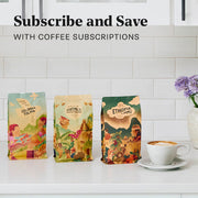 Three colorful Whole Latte Love coffee bean bags labeled Colombia Tolima, Guatemala Huehuetenango, and Ethiopia Limmu sit on a white countertop beside a latte, with a white subway-tile wall behind them and the text 'Subscribe and Save WITH COFFEE SUBSCRIPTIONS' overlayed at the top.