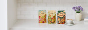 Three bags of Whole Latte Love coffee with colorful illustrated packaging stand on a white countertop against a white subway tile backsplash. A white latte cup and saucer with coffee sit to the right, next to a small vase of purple flowers.