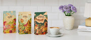 Close-up of three bags of Whole Latte Love coffee with colorful illustrated packaging stand on a white countertop against a white subway tile backsplash. A white latte cup and saucer with coffee sit to the right, next to a small vase of purple flowers.