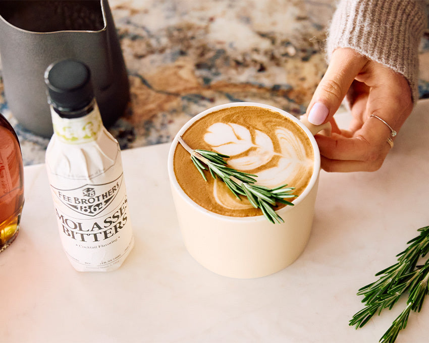 Cup of coffee with latte art and rosemary garnish, accompanied by a bottle of molasses bitter on a marble surface.
